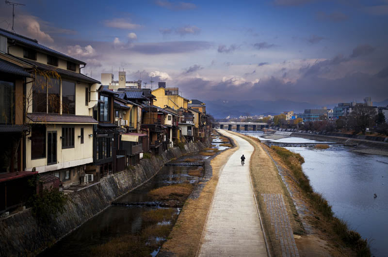 A scenic view of a riverbank pathway lined with traditional Japanese houses on the left, under a cloudy blue sky. A small cyclist travels along the path beside the calm river.